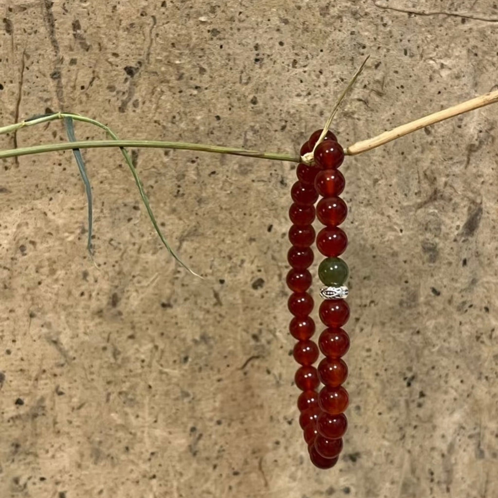 A handcrafted CIRCLEARA He Huan bracelet with 6mm red agate and a single green jade bead, styled in a minimalist earthen setting.