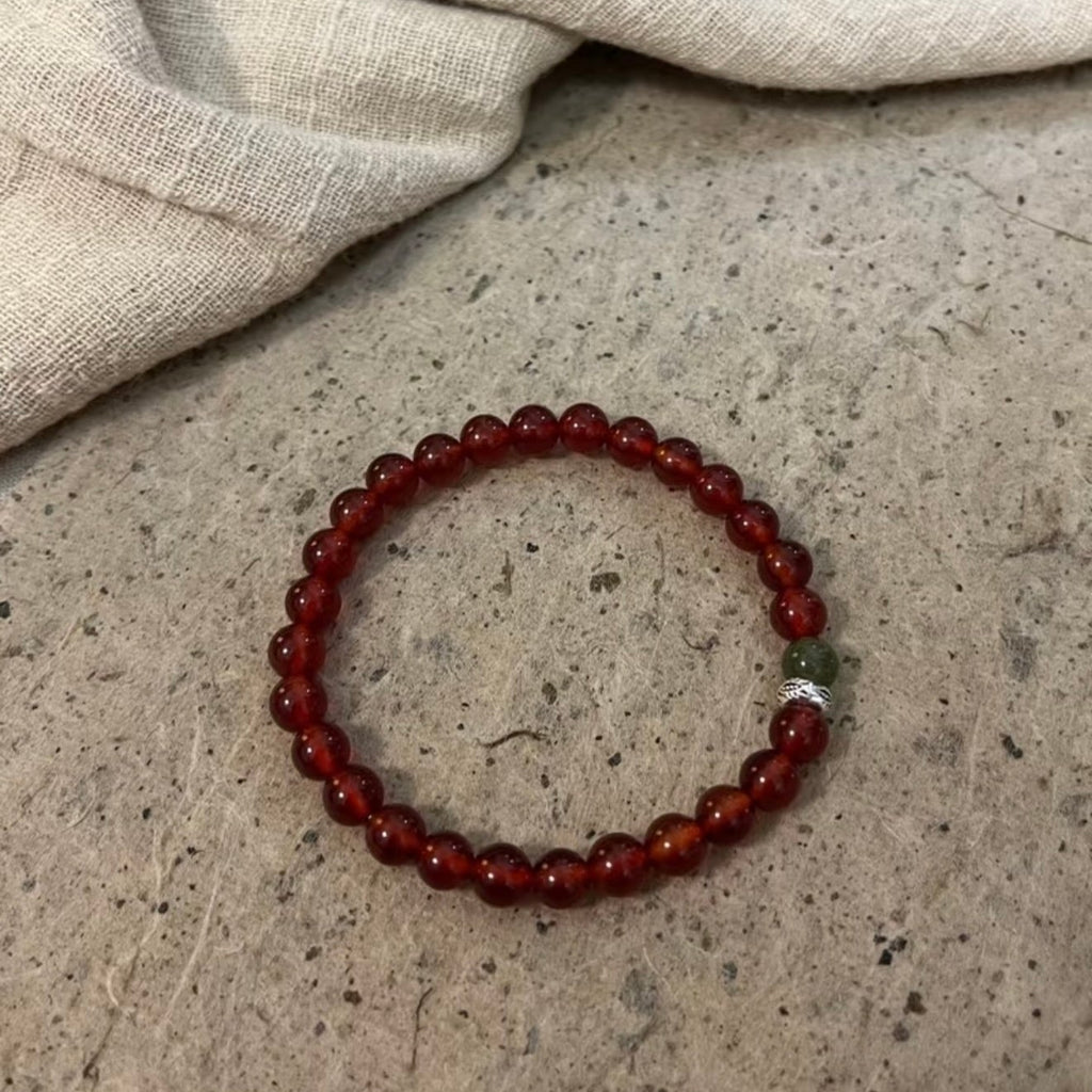 A handcrafted CIRCLEARA He Huan bracelet with 6mm red agate and a single green jade bead, styled in a minimalist earthen setting.