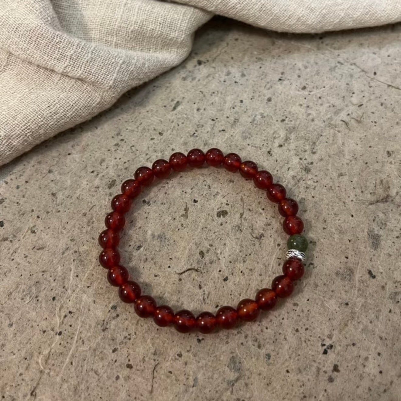 A handcrafted CIRCLEARA He Huan bracelet with 6mm red agate and a single green jade bead, styled in a minimalist earthen setting.