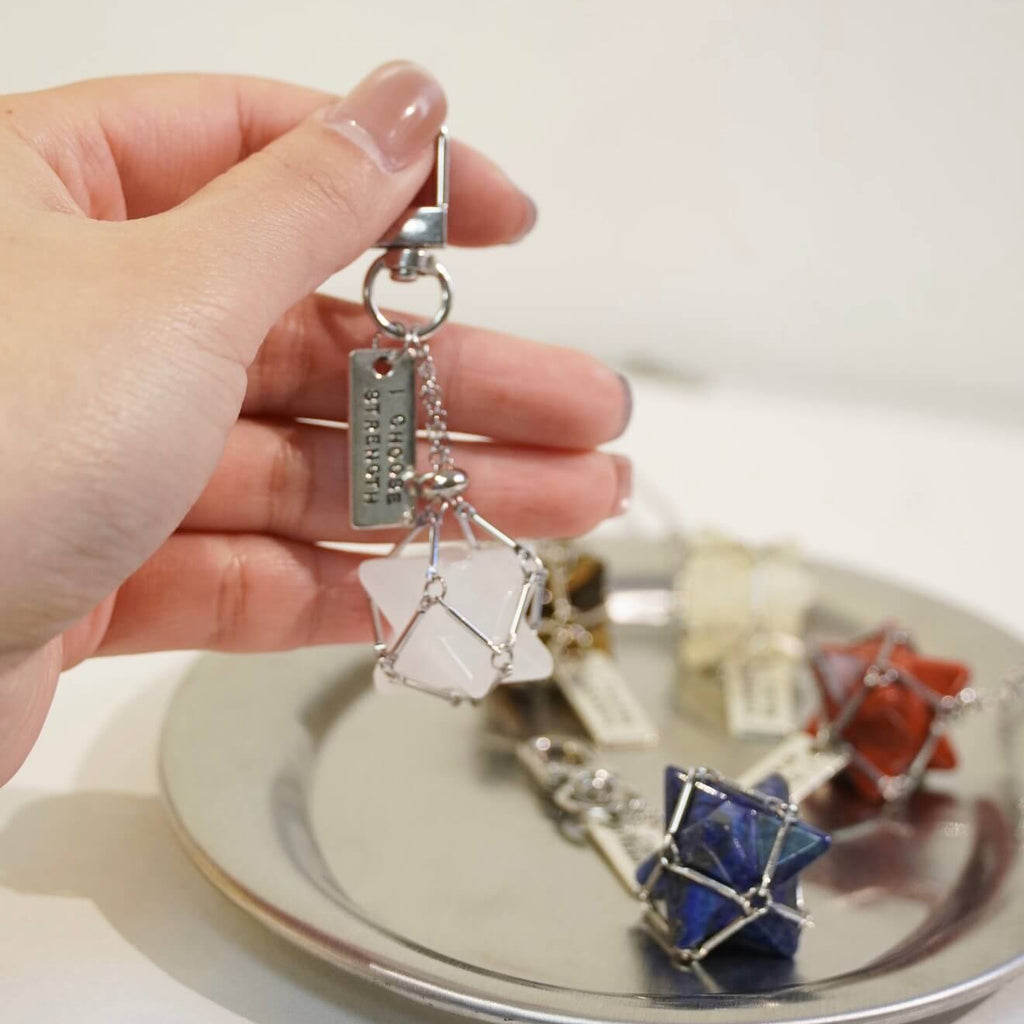 Handcrafted Red Jasper Merkaba star sacred geometry crystal accessory, featuring a protective alloy cage, heavy-duty keyring clip, and motivational tag on a minimalist background.