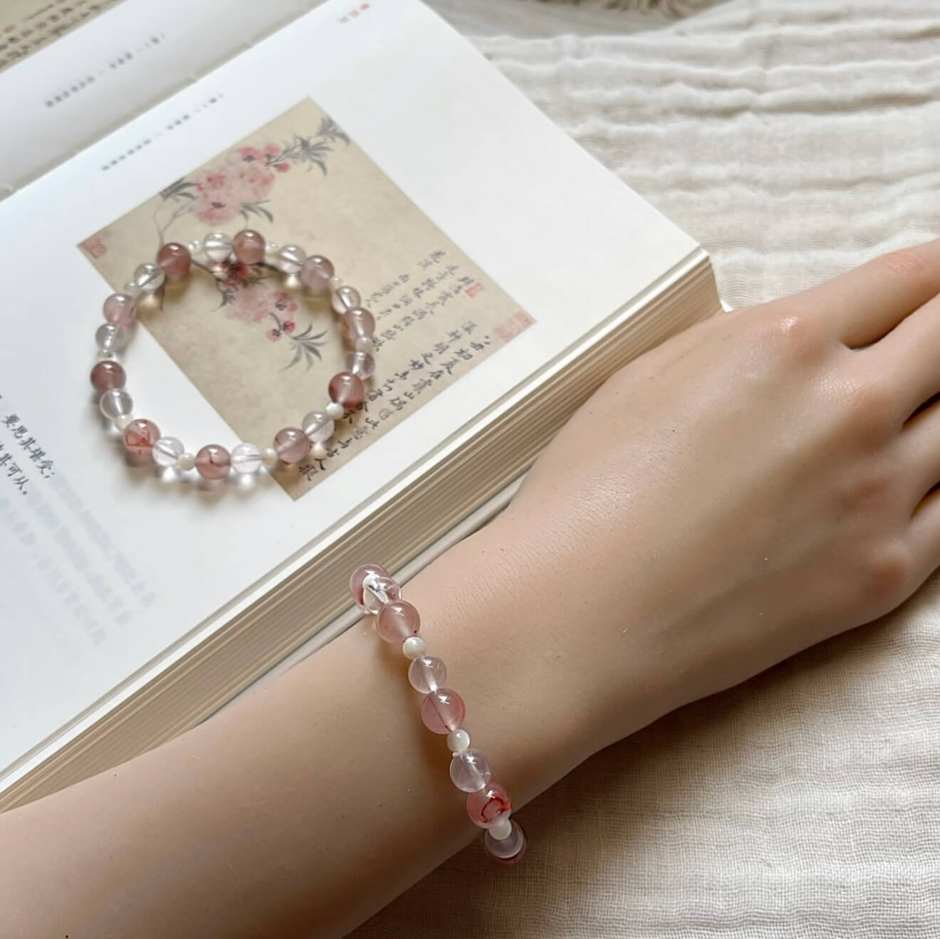 Two handcrafted Rose Quartz and Southern Red Agate grounding bracelets with white shell accents, displayed on a textured linen cloth alongside an artistic vintage book.