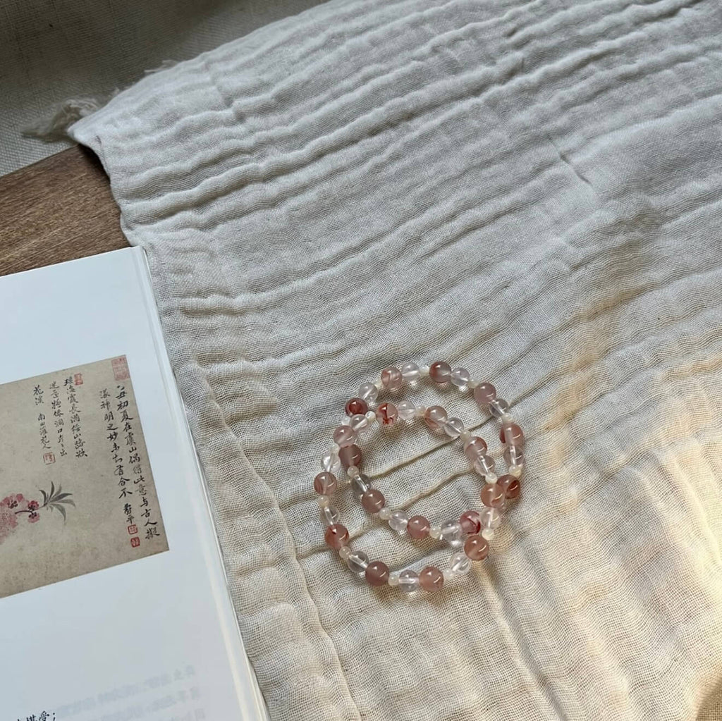 Two handcrafted Rose Quartz and Southern Red Agate grounding bracelets with white shell accents, displayed on a textured linen cloth alongside an artistic vintage book.