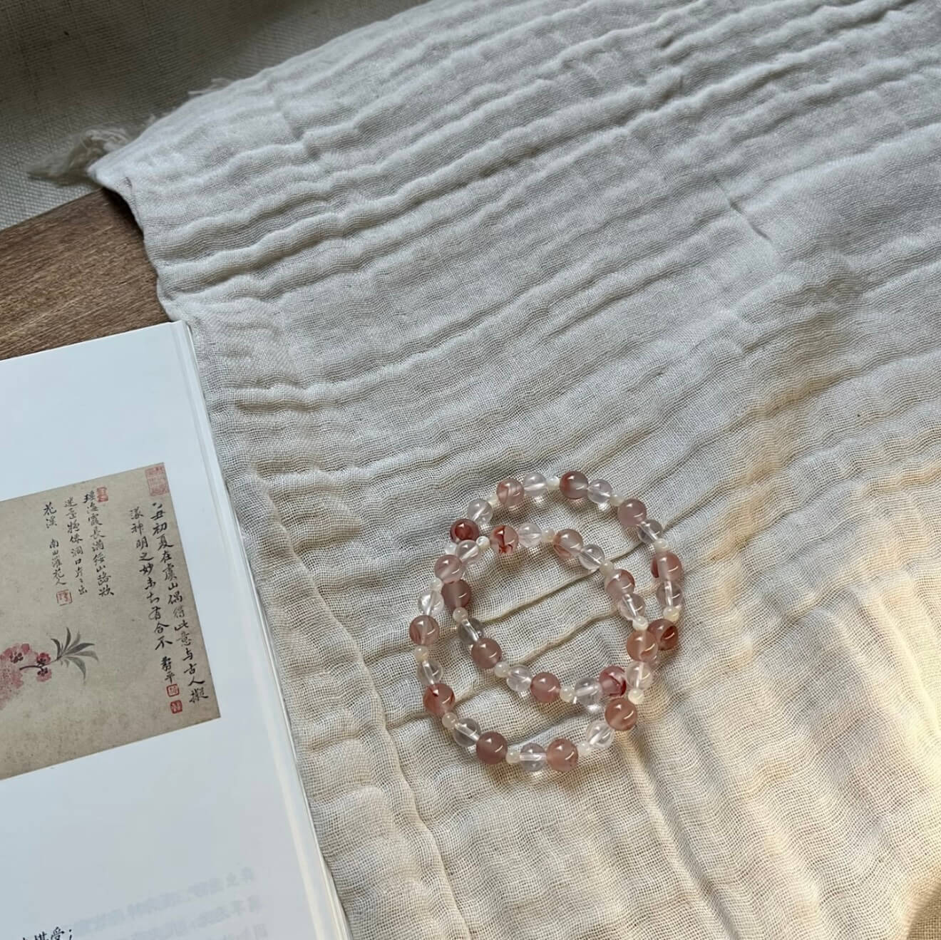 Two handcrafted Rose Quartz and Southern Red Agate grounding bracelets with white shell accents, displayed on a textured linen cloth alongside an artistic vintage book.