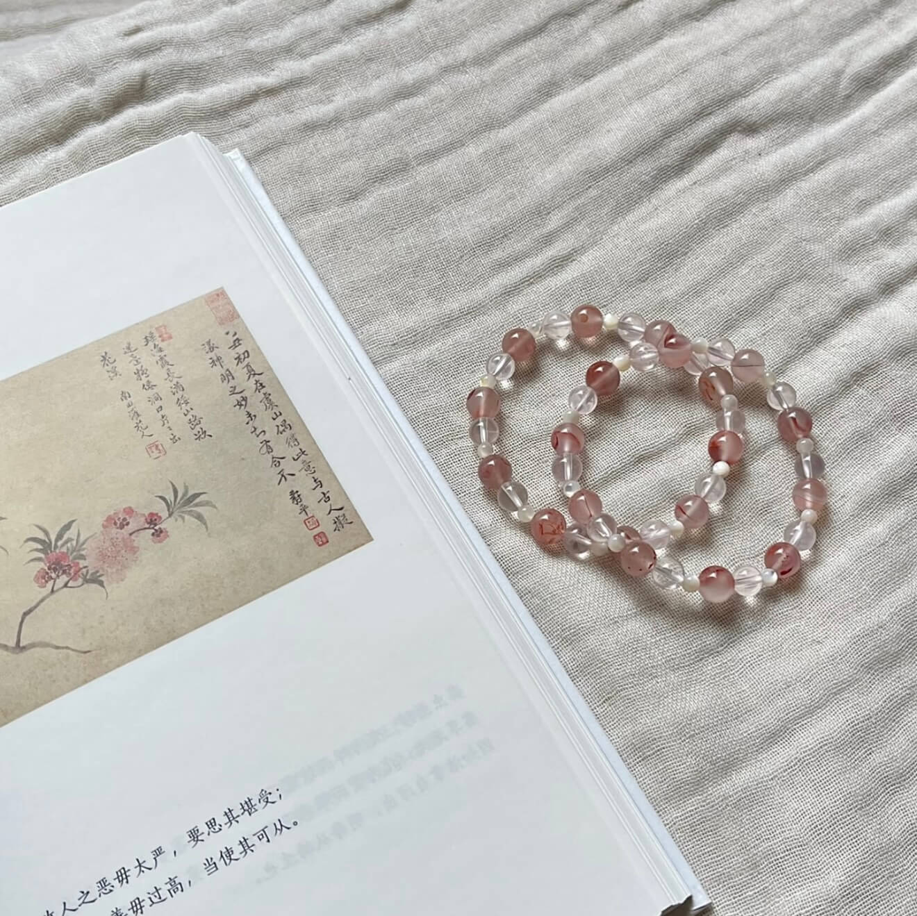 Two handcrafted Rose Quartz and Southern Red Agate grounding bracelets with white shell accents, displayed on a textured linen cloth alongside an artistic vintage book.