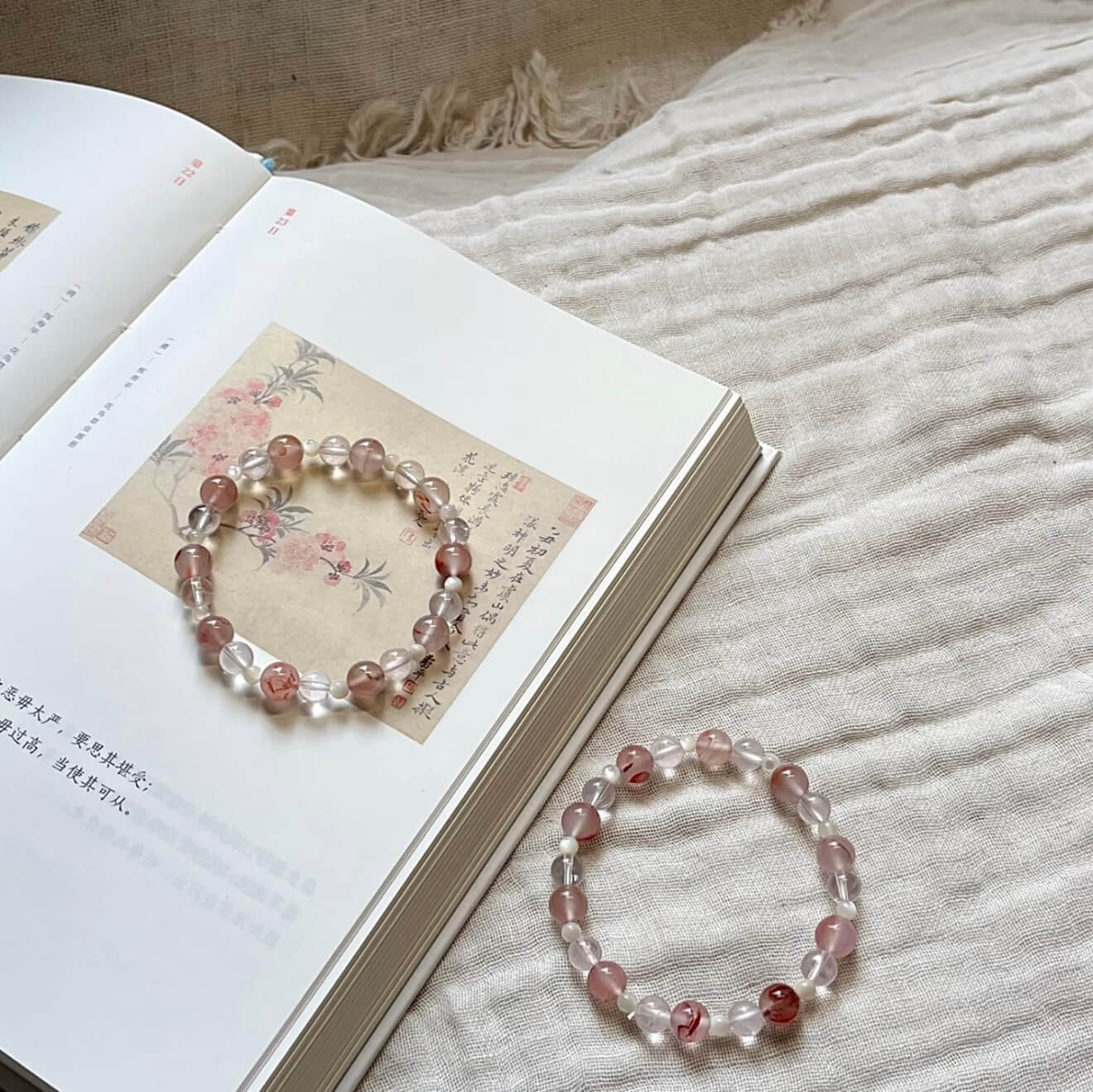 Two handcrafted Rose Quartz and Southern Red Agate grounding bracelets with white shell accents, displayed on a textured linen cloth alongside an artistic vintage book.