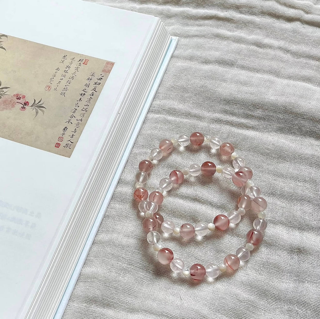 Two handcrafted Rose Quartz and Southern Red Agate grounding bracelets with white shell accents, displayed on a textured linen cloth alongside an artistic vintage book.