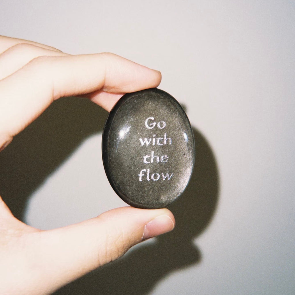 Handcrafted natural dark mystic stone with silver-etched inscription 'Go with the flow' by CIRCLEARA, resting on a minimalist white ceramic display holder.