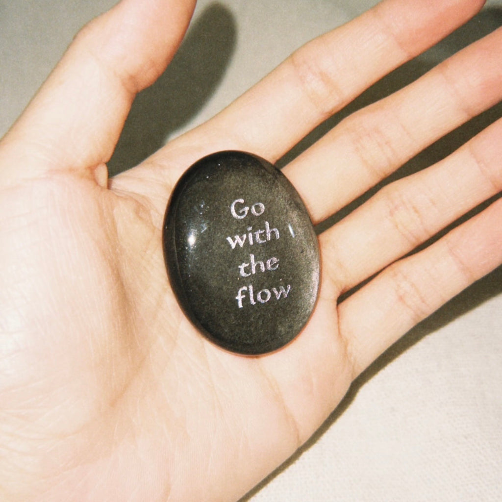 Handcrafted natural dark mystic stone with silver-etched inscription 'Go with the flow' by CIRCLEARA, resting on a minimalist white ceramic display holder.