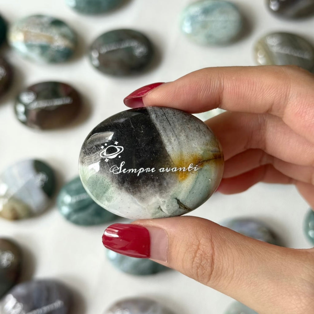 Handcrafted 45mm natural Ocean Jasper cosmic palm stones by CIRCLEARA, engraved with silver text 'KEEP MOVING' and a starburst symbol, displaying unique green and white orbicular textures.