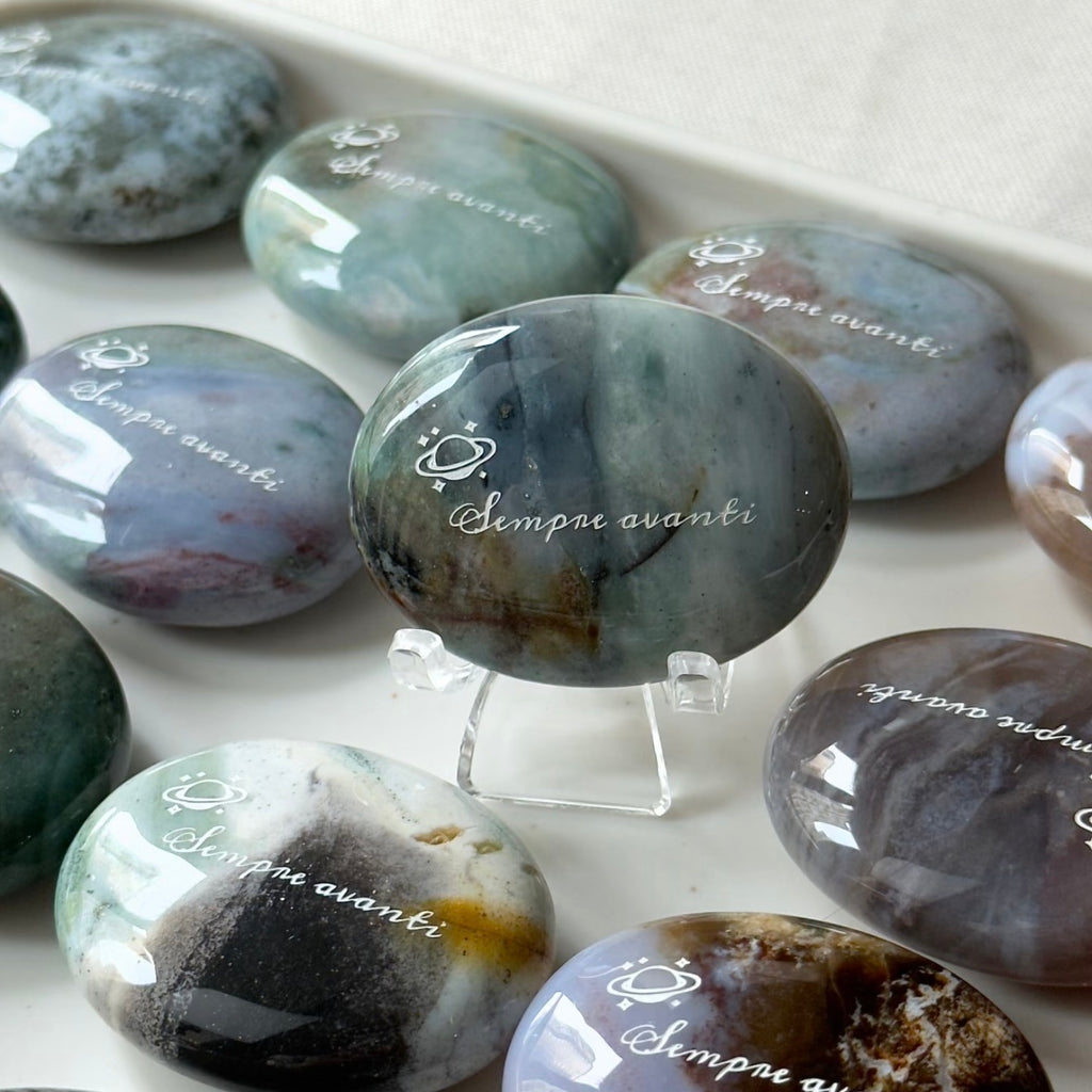 Handcrafted 45mm natural Ocean Jasper cosmic palm stones by CIRCLEARA, engraved with silver text 'KEEP MOVING' and a starburst symbol, displaying unique green and white orbicular textures.