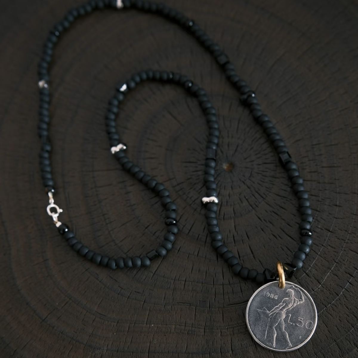 Close-up shot of CIRCLEARA boundary necklace featuring a weighted silver vintage coin pendant with an engraved classical deity, connected by a brass bail to premium Japanese Miyuki matte black glass beads.