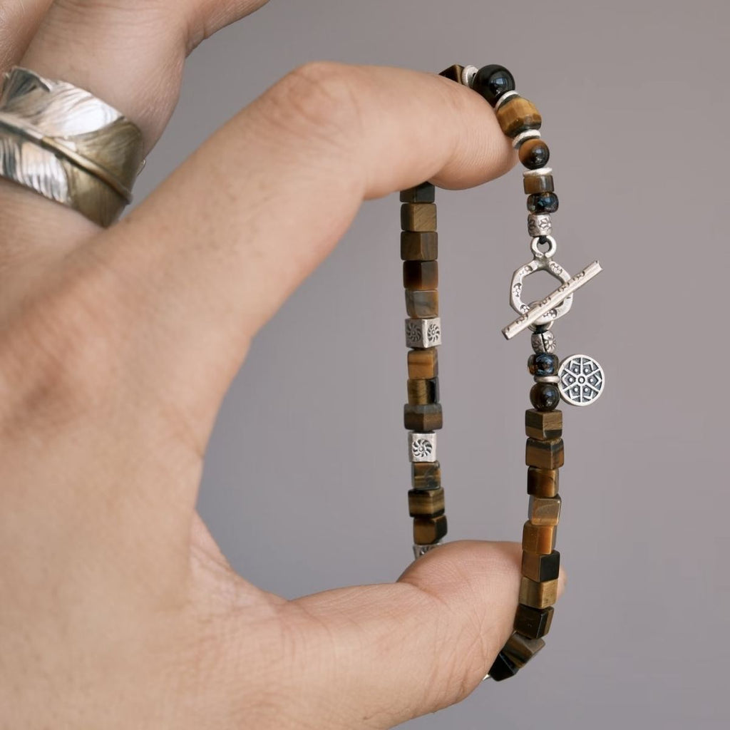A flat-lay layout displaying the CIRCLEARA focus bracelet composed of geometric cube-cut brown tiger's eye stones, hand-hammered 925 sterling silver spacers, and an intricate circular silver charm, showing an organic minimalist style.