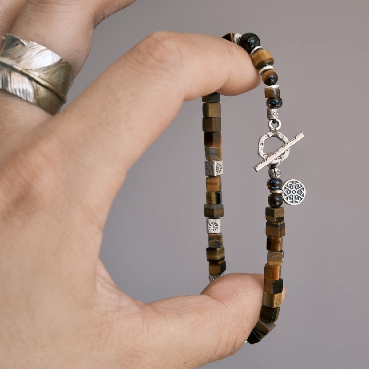 A flat-lay layout displaying the CIRCLEARA focus bracelet composed of geometric cube-cut brown tiger's eye stones, hand-hammered 925 sterling silver spacers, and an intricate circular silver charm, showing an organic minimalist style.