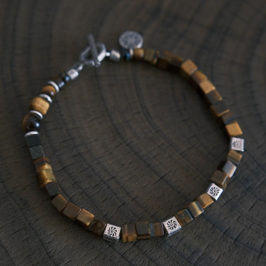 A flat-lay layout displaying the CIRCLEARA focus bracelet composed of geometric cube-cut brown tiger's eye stones, hand-hammered 925 sterling silver spacers, and an intricate circular silver charm, showing an organic minimalist style.