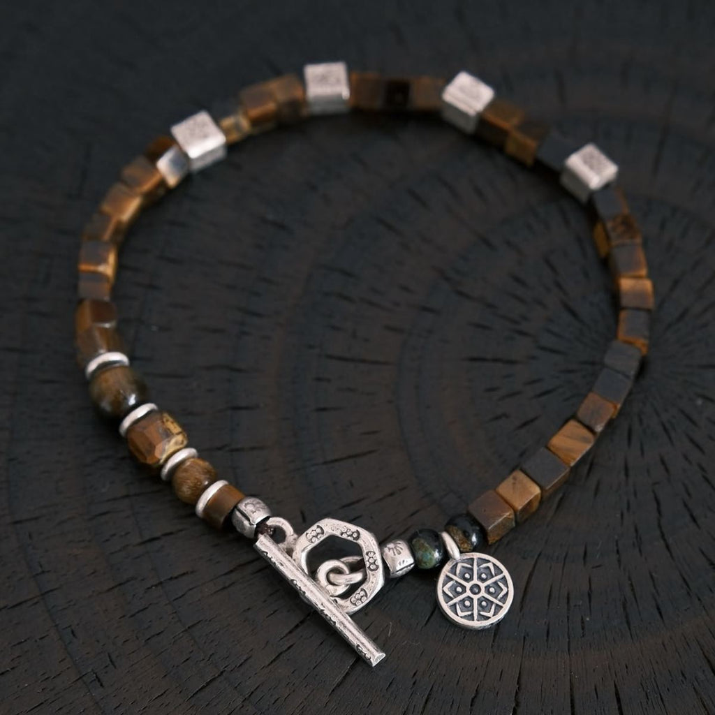 A flat-lay layout displaying the CIRCLEARA focus bracelet composed of geometric cube-cut brown tiger's eye stones, hand-hammered 925 sterling silver spacers, and an intricate circular silver charm, showing an organic minimalist style.