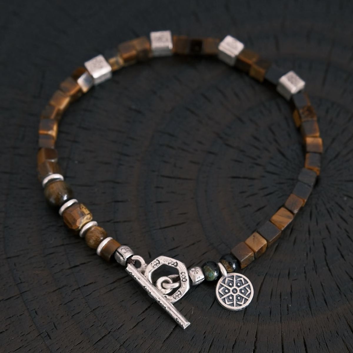 A flat-lay layout displaying the CIRCLEARA focus bracelet composed of geometric cube-cut brown tiger's eye stones, hand-hammered 925 sterling silver spacers, and an intricate circular silver charm, showing an organic minimalist style.