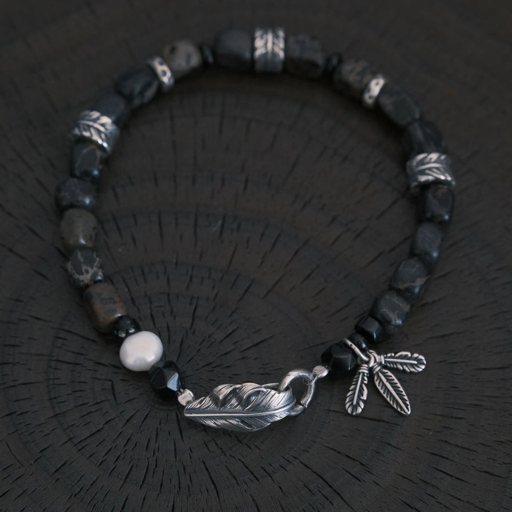 A high-contrast vertical shot of the CIRCLEARA Winged Sovereign bracelet, highlighting the detailed sequence of natural grey-brown veined jasper cubes, faceted black onyx, a single white baroque pearl, and an intricate 925 sterling silver feather charm.