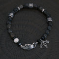 A high-contrast vertical shot of the CIRCLEARA Winged Sovereign bracelet, highlighting the detailed sequence of natural grey-brown veined jasper cubes, faceted black onyx, a single white baroque pearl, and an intricate 925 sterling silver feather charm.
