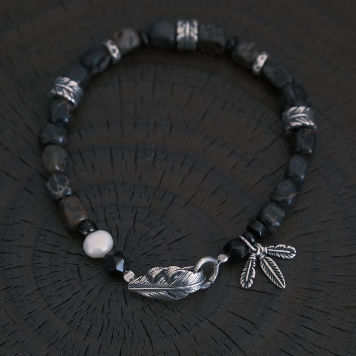 A high-contrast vertical shot of the CIRCLEARA Winged Sovereign bracelet, highlighting the detailed sequence of natural grey-brown veined jasper cubes, faceted black onyx, a single white baroque pearl, and an intricate 925 sterling silver feather charm.