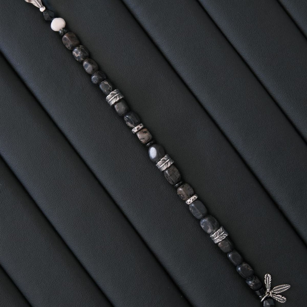 A high-contrast vertical shot of the CIRCLEARA Winged Sovereign bracelet, highlighting the detailed sequence of natural grey-brown veined jasper cubes, faceted black onyx, a single white baroque pearl, and an intricate 925 sterling silver feather charm.