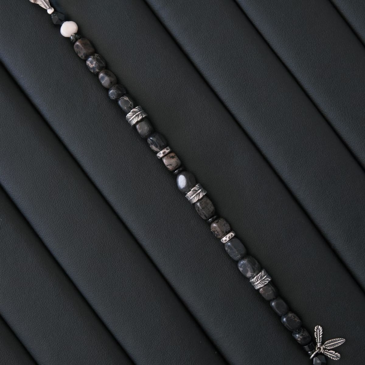 A high-contrast vertical shot of the CIRCLEARA Winged Sovereign bracelet, highlighting the detailed sequence of natural grey-brown veined jasper cubes, faceted black onyx, a single white baroque pearl, and an intricate 925 sterling silver feather charm.