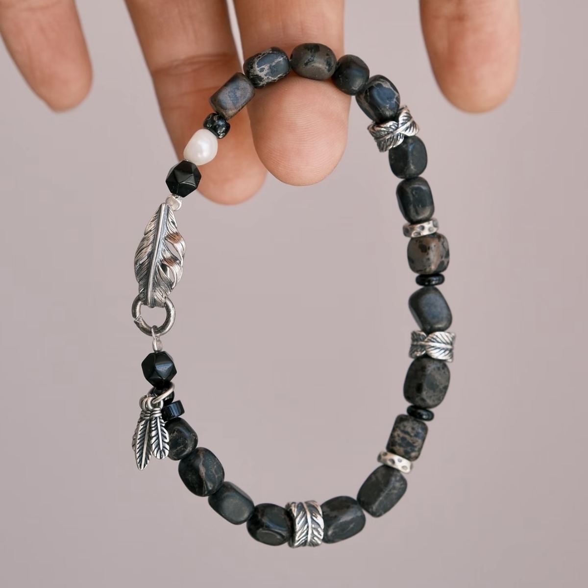 A high-contrast vertical shot of the CIRCLEARA Winged Sovereign bracelet, highlighting the detailed sequence of natural grey-brown veined jasper cubes, faceted black onyx, a single white baroque pearl, and an intricate 925 sterling silver feather charm.