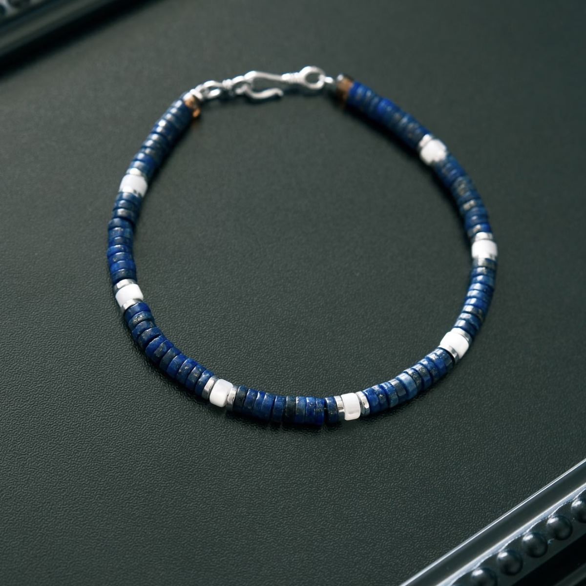 A modern minimalist lifestyle photo of the unisex CIRCLEARA lapis lazuli and silver beaded bracelet styled on a model's wrist, layered alongside a silver chain ring, highlighting a quiet luxury and stoic aesthetic.