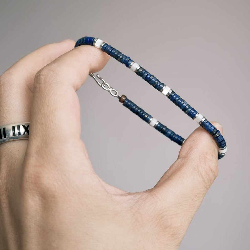 A modern minimalist lifestyle photo of the unisex CIRCLEARA lapis lazuli and silver beaded bracelet styled on a model's wrist, layered alongside a silver chain ring, highlighting a quiet luxury and stoic aesthetic.