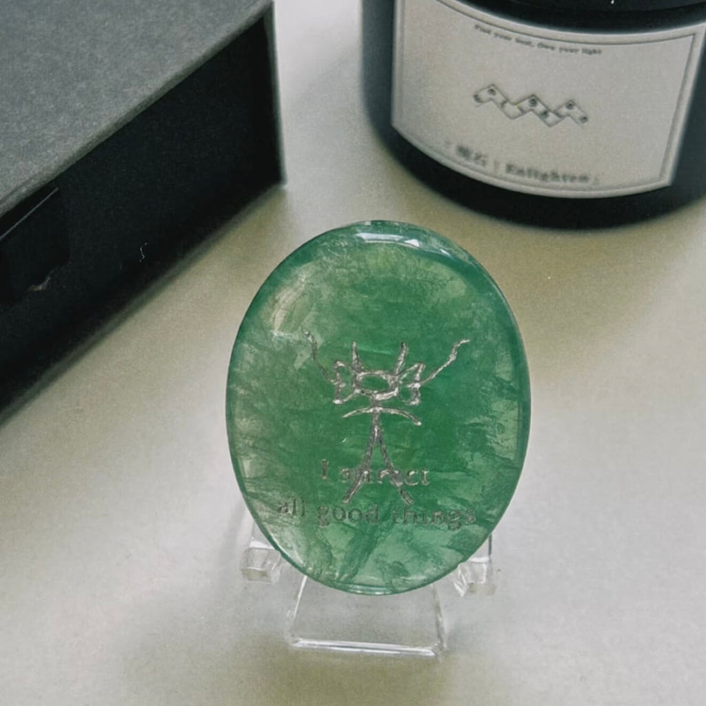 A front-facing detailed shot of the translucent green fluorite CIRCLEARA palm stone resting on its complimentary clear minimalist stand, revealing the precise Dongba pictograph and the etched phrase "I attract all good things."