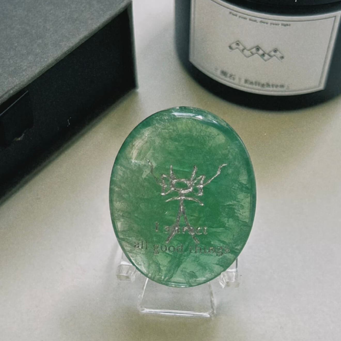A front-facing detailed shot of the translucent green fluorite CIRCLEARA palm stone resting on its complimentary clear minimalist stand, revealing the precise Dongba pictograph and the etched phrase "I attract all good things."