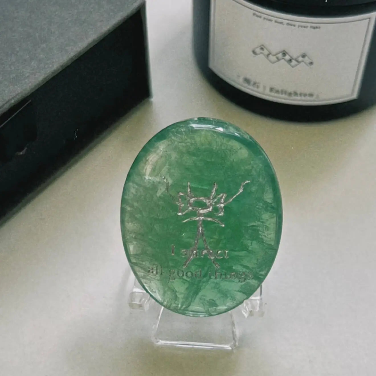 A front-facing detailed shot of the translucent green fluorite CIRCLEARA palm stone resting on its complimentary clear minimalist stand, revealing the precise Dongba pictograph and the etched phrase "I attract all good things."