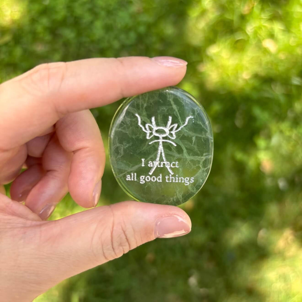 A front-facing detailed shot of the translucent green fluorite CIRCLEARA palm stone resting on its complimentary clear minimalist stand, revealing the precise Dongba pictograph and the etched phrase "I attract all good things."