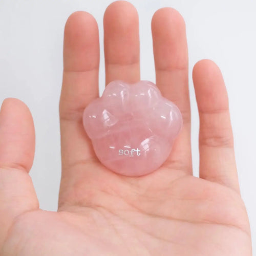 A close-up shot of a hand gently cradling the smooth, translucent pink rose quartz CIRCLEARA palm stone shaped like a rounded cat paw pad with the etched text "soft".
