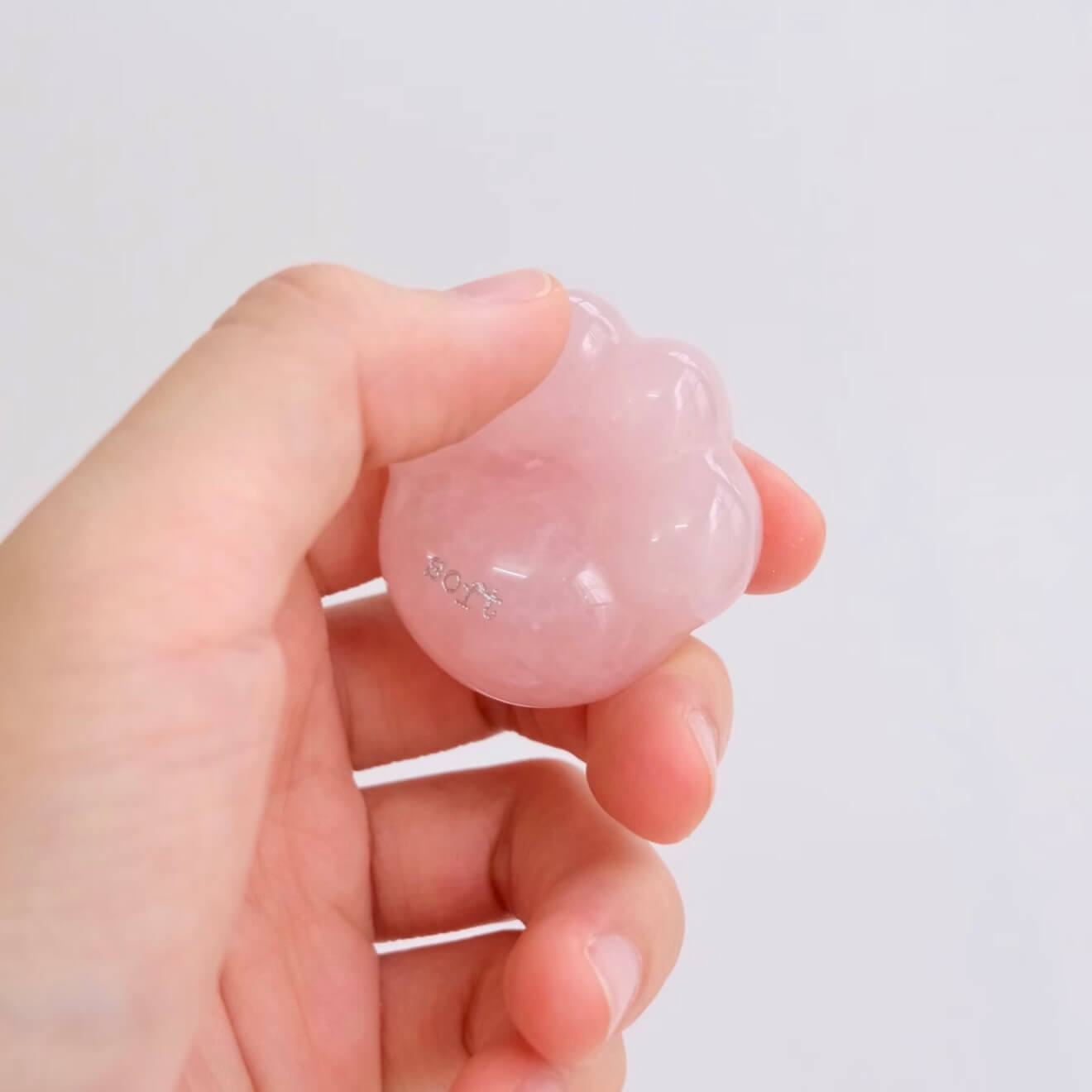 A close-up shot of a hand gently cradling the smooth, translucent pink rose quartz CIRCLEARA palm stone shaped like a rounded cat paw pad with the etched text "soft".
