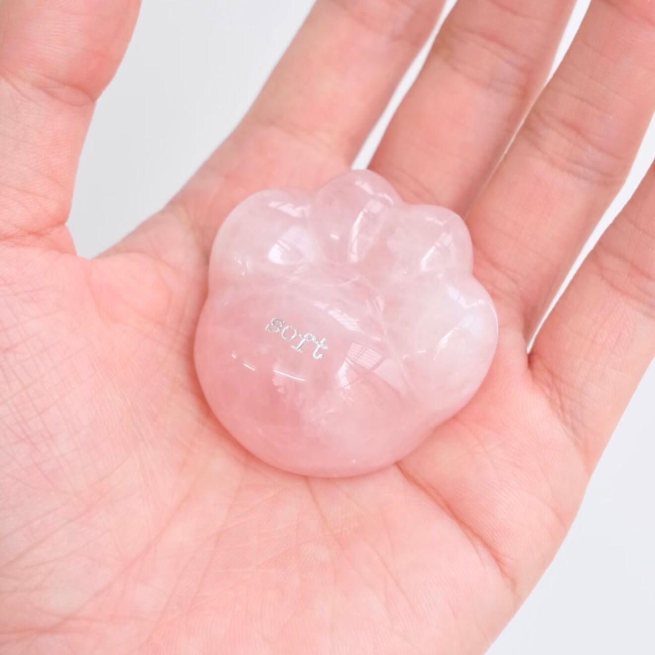 A close-up shot of a hand gently cradling the smooth, translucent pink rose quartz CIRCLEARA palm stone shaped like a rounded cat paw pad with the etched text "soft".