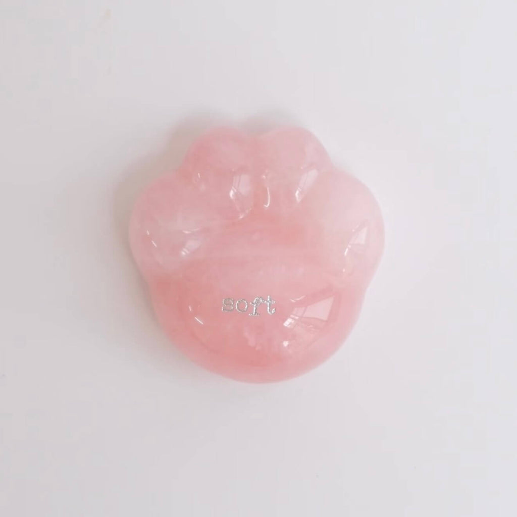 A close-up shot of a hand gently cradling the smooth, translucent pink rose quartz CIRCLEARA palm stone shaped like a rounded cat paw pad with the etched text "soft".