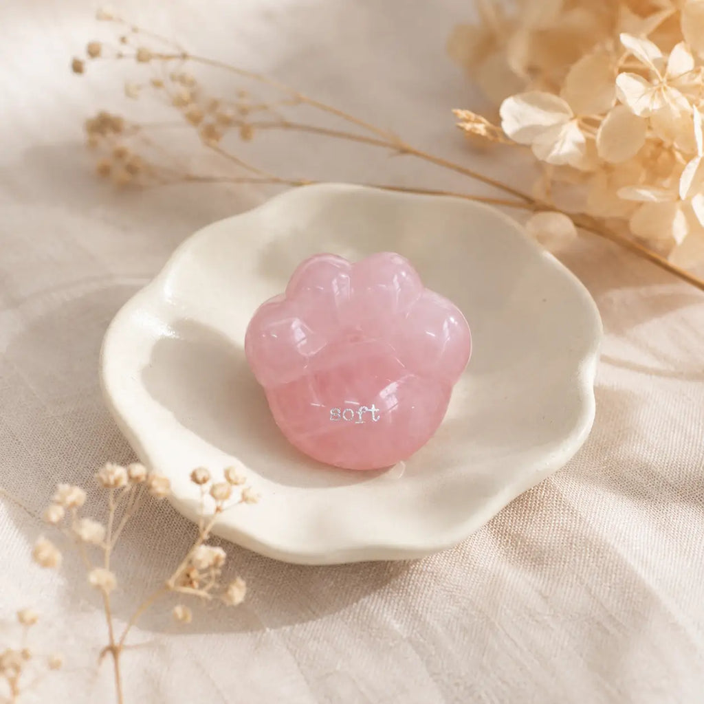 A close-up shot of a hand gently cradling the smooth, translucent pink rose quartz CIRCLEARA palm stone shaped like a rounded cat paw pad with the etched text "soft".