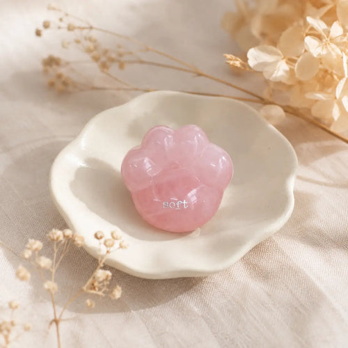 A close-up shot of a hand gently cradling the smooth, translucent pink rose quartz CIRCLEARA palm stone shaped like a rounded cat paw pad with the etched text "soft".