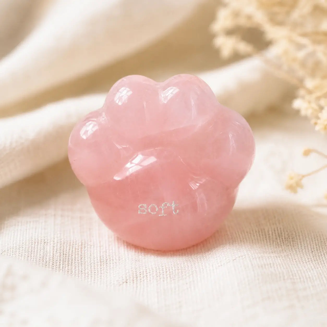A close-up shot of a hand gently cradling the smooth, translucent pink rose quartz CIRCLEARA palm stone shaped like a rounded cat paw pad with the etched text "soft".
