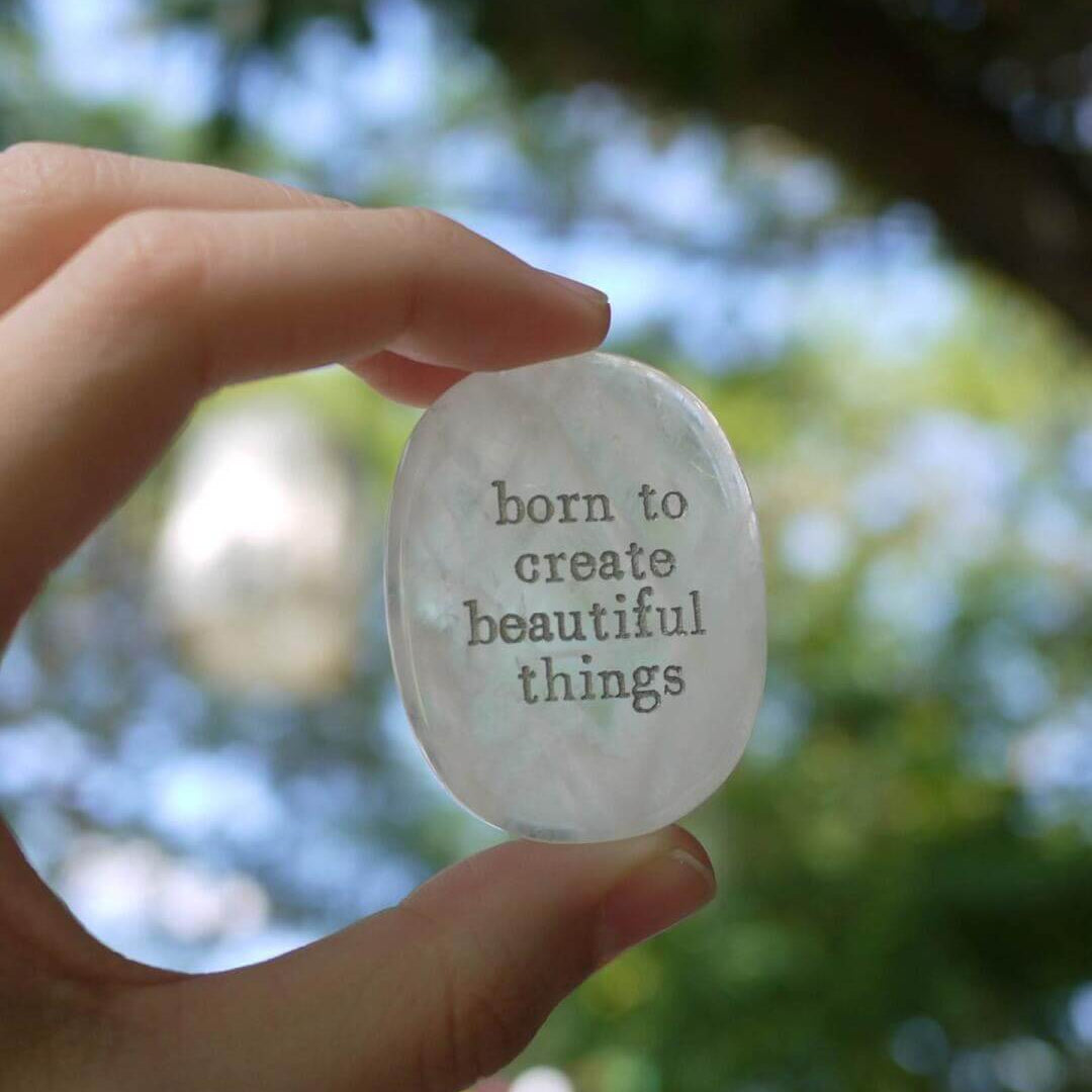 Hand holding a CIRCLEARA etched affirmation crystal stone, translucent texture, "born to create beautiful things" creative manifestation tool.