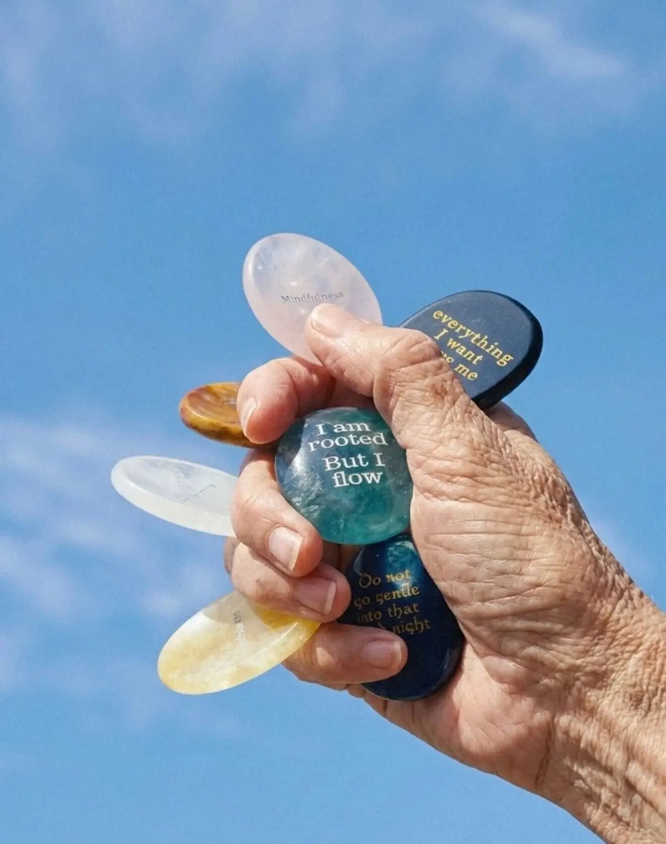 A hand holding several crystal affirmation stones against a blue sky background.